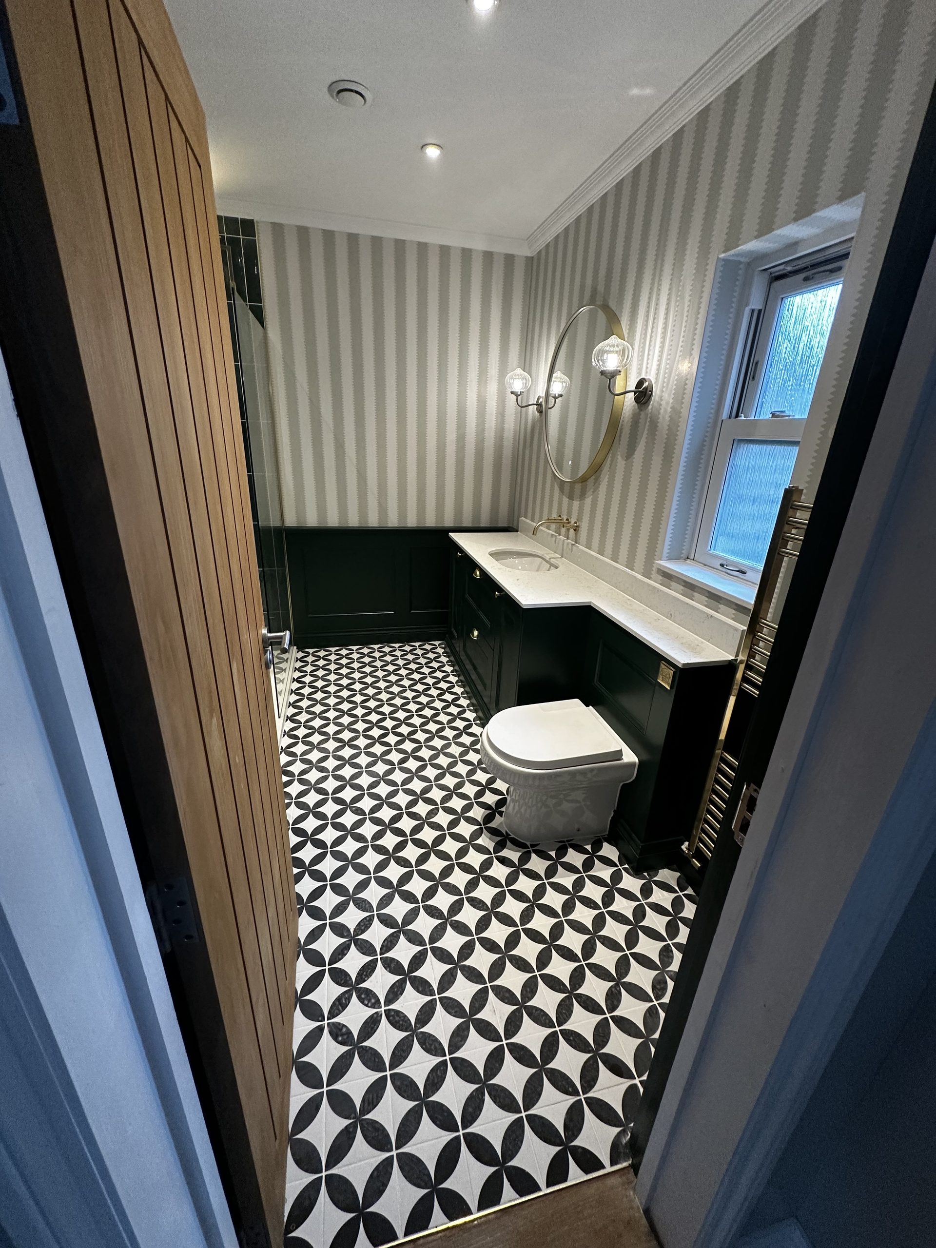 Cloakroom with dark green panelling and vanity unit, striped wallpaper, patterned monochrome floor tiles and a round mirror