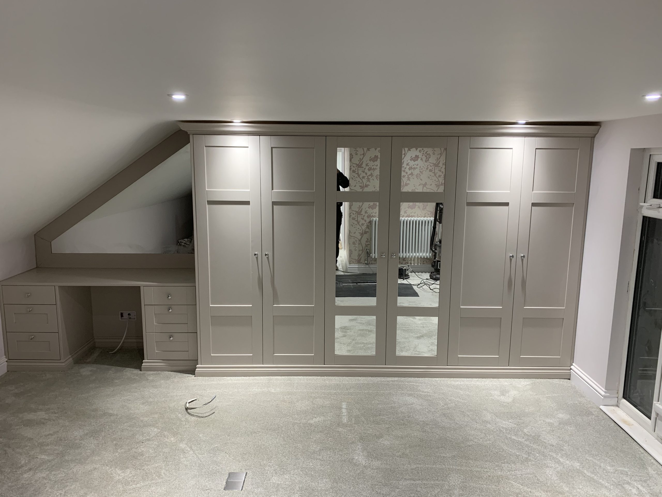 Fitted wardrobe with taupe shaker doors and mirrored centre panels, built into a loft bedroom beside a matching desk under the eaves