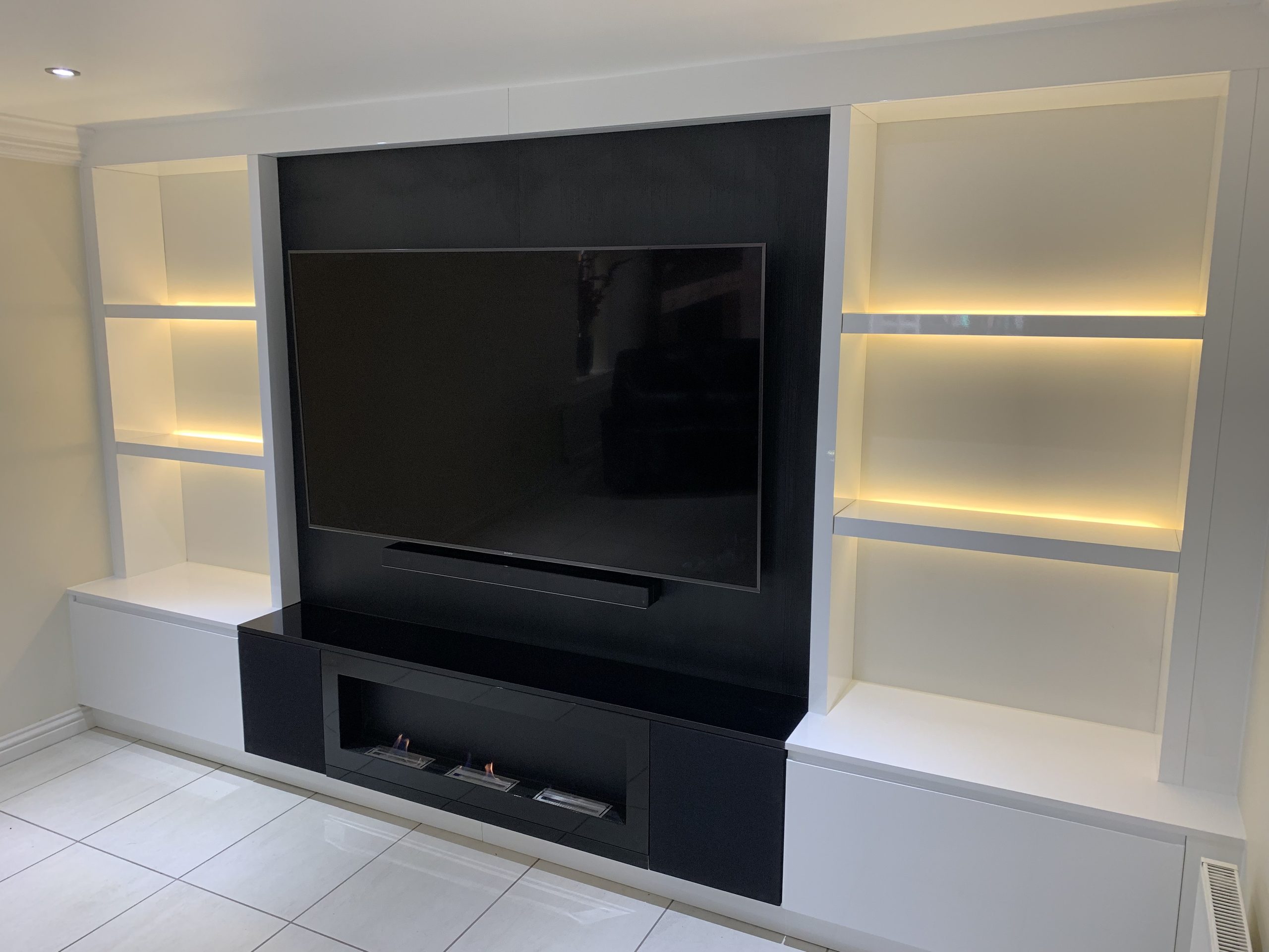 Modern media wall with a wall mounted TV on a dark panel, flanked by white LED lit display shelving above a linear fireplace